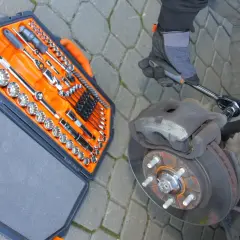 Ręka w rękawicy odkręca zapieczoną śrubę przy hamulcu. Obok walizka z narzędziami.