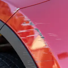 Porysowane auto z czerwonym lakierem, widoczne głębokie rysy i otarcia na błotniku, obok czarna opona i plastikowa osłona nadkola.