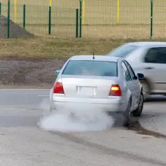 Srebrny samochód z silnikiem diesla kopci na biało, tworząc gęstą chmurę dymu za sobą.