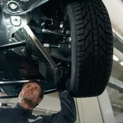 Mechanik bada podwozie auta, szukając przyczyny stukania w kole. Używa latarki, by lepiej widzieć.