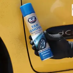 Rękawiczka z czarną gumą trzyma puszkę K2 Tar Remover, która skutecznie usuwa smołę z żółtego lakieru samochodu.