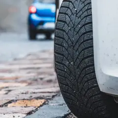 Białe auto z zimową oponą na brukowanej ulicy. Czy można wymienić jedną oponę? W tle niebieski samochód.