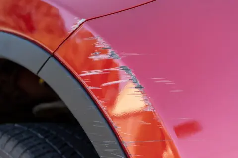 Porysowane auto z czerwonym lakierem, widoczne głębokie rysy i otarcia na błotniku, obok czarna opona i plastikowa osłona nadkola.