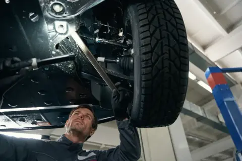 Mechanik bada podwozie auta, szukając przyczyny stukania w kole. Używa latarki, by lepiej widzieć.