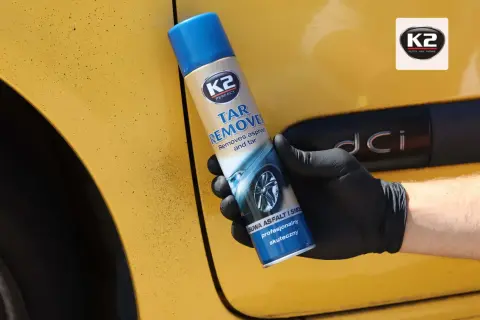 Rękawiczka z czarną gumą trzyma puszkę K2 Tar Remover, która skutecznie usuwa smołę z żółtego lakieru samochodu.
