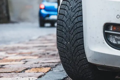 Białe auto z zimową oponą na brukowanej ulicy. Czy można wymienić jedną oponę? W tle niebieski samochód.