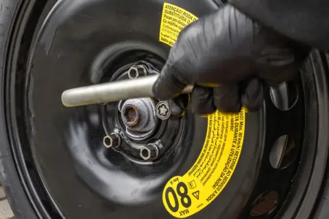 Ręka w czarnej rękawiczce dokręca śrubę koła dojazdowego. Na naklejce widnieje informacja o maksymalnej prędkości 80 km/h, ale nie podano jakie ciśnienie w kole dojazdowym jest zalecane.