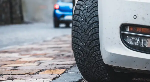 Białe auto z zimową oponą na brukowanej ulicy. Czy można wymienić jedną oponę? W tle niebieski samochód.