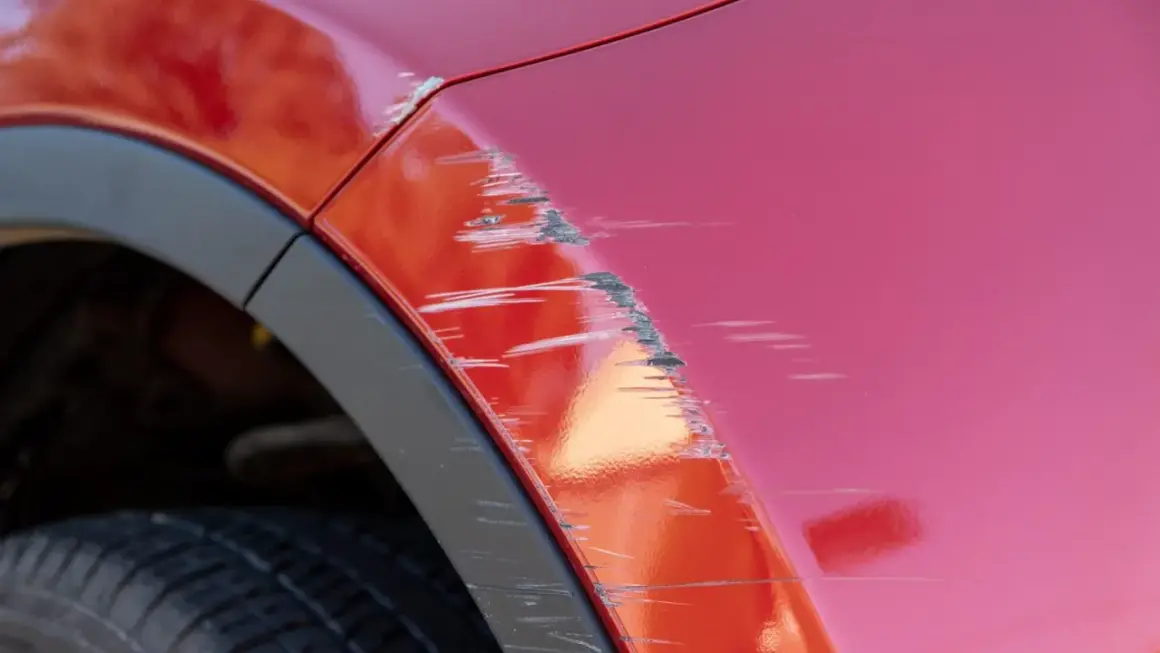 Porysowane auto z czerwonym lakierem, widoczne głębokie rysy i otarcia na błotniku, obok czarna opona i plastikowa osłona nadkola.