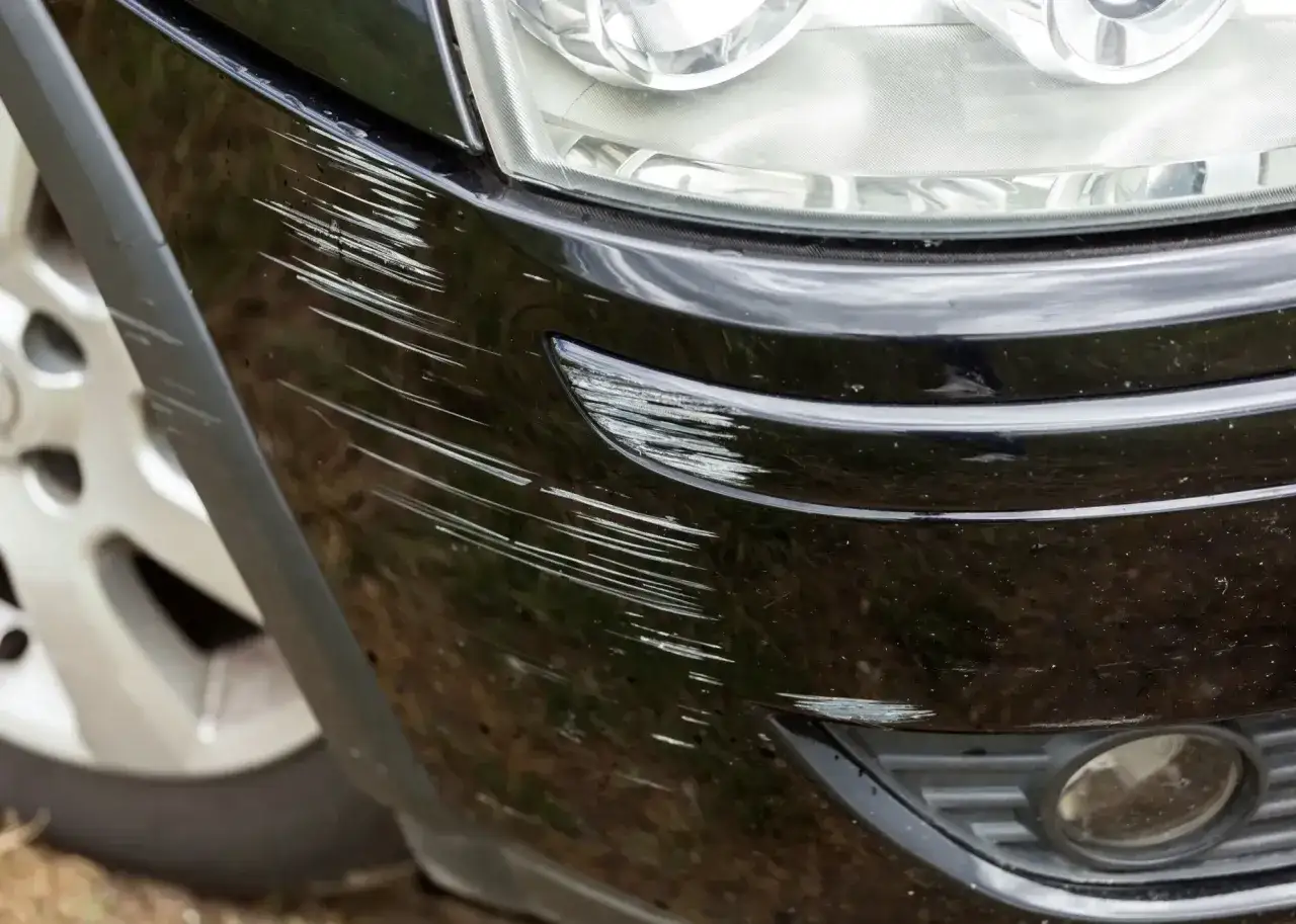 Porysowane auto z licznymi zadrapaniami na czarnym zderzaku i błotniku. Widać fragment reflektora i felgi.
