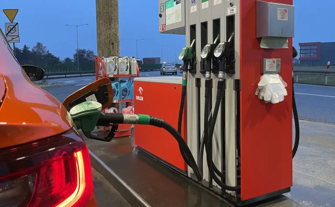 Tankowanie pomarańczowego auta na stacji Orlen. Dostępna benzyna E10, widać dystrybutory i akcesoria.