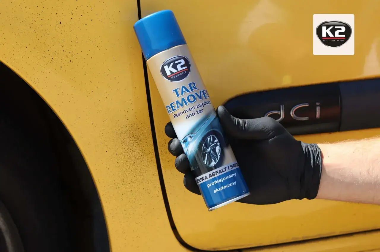 Rękawiczka z czarną gumką trzyma puszkę K2 Tar Remover, kt&oacute;ra skutecznie usuwa smołę z ż&oacute;łtego lakieru samochodu.