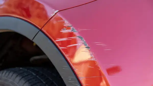 Porysowane auto z czerwonym lakierem, widoczne głębokie rysy i otarcia na błotniku, obok czarna opona i plastikowa osłona nadkola.