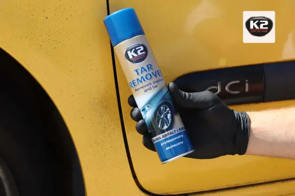 Rękawiczka z czarną gumą trzyma puszkę K2 Tar Remover, która skutecznie usuwa smołę z żółtego lakieru samochodu.